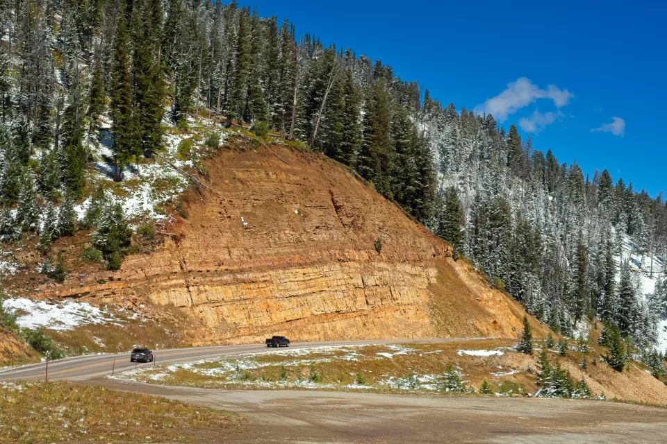 A road winds along the edge of a mountain, with an inclined tan rockface above it and a snowy treeline near the top of a photo.